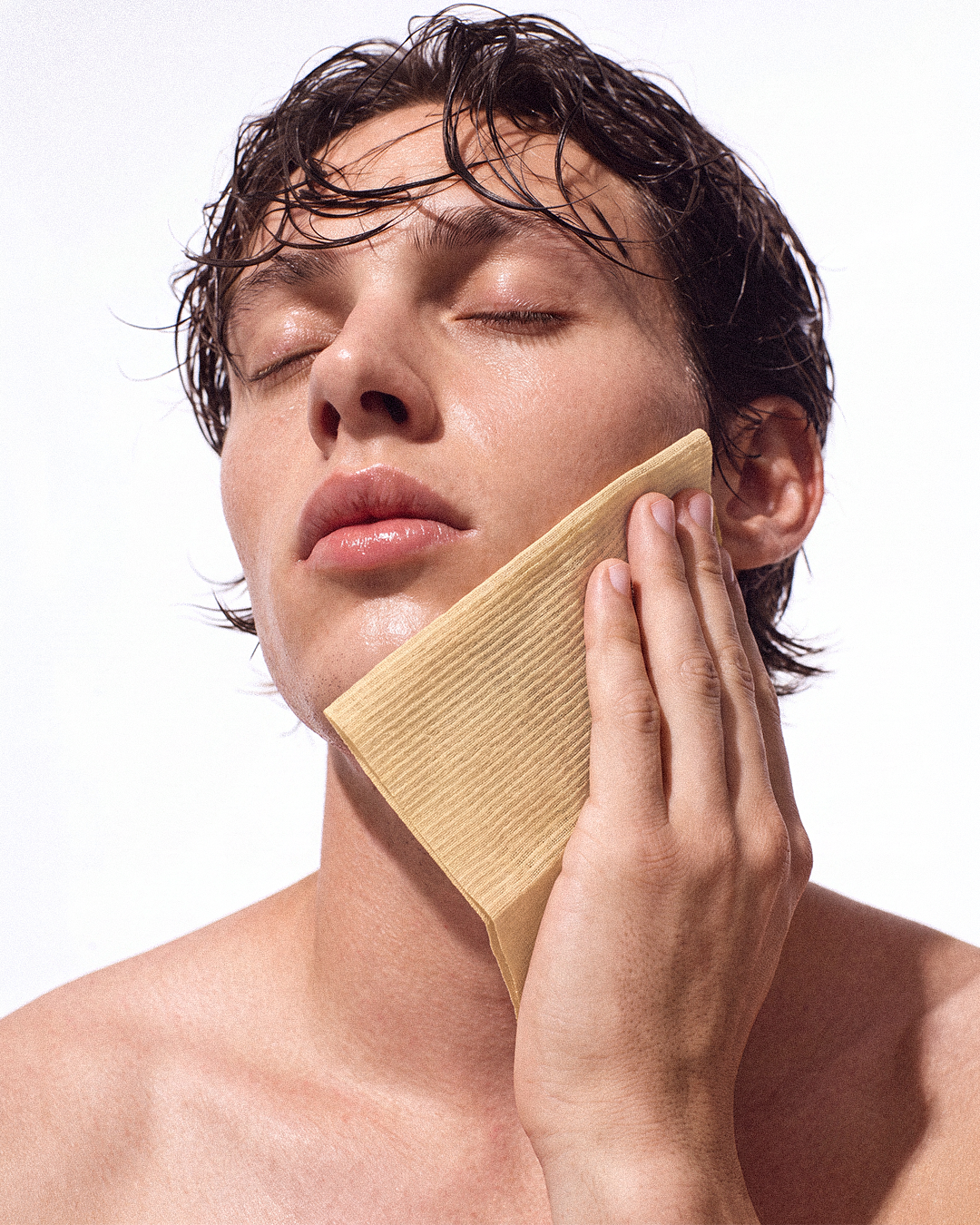 A close-up of a man with wet dark hair holding a single use face towel against his cheek, with his eyes closed, against a plain white background.