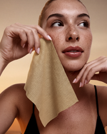 A close-up lifestyle shot of a woman with freckles holding a dual-sided single use beige facial cleansing towel against her cheek.