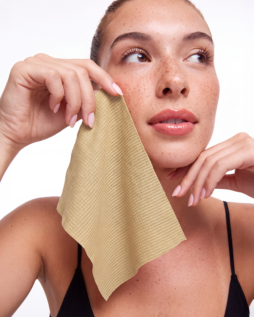 A woman with freckles holds a single use face towel near her face against a clean white background.